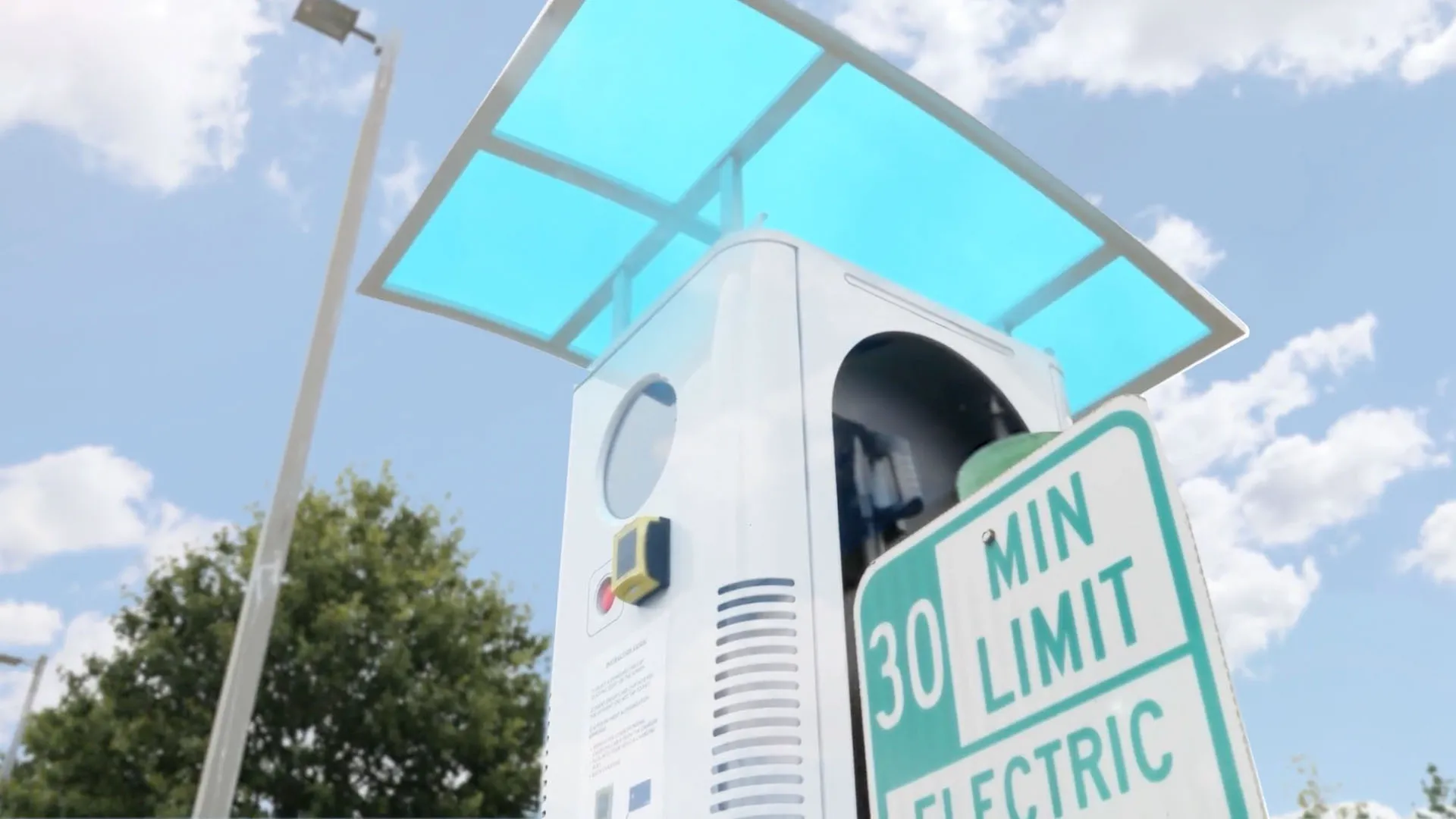 An electric vehicle fast charger under a blue canopy, beside a road sign reading "30 min limit, electric".